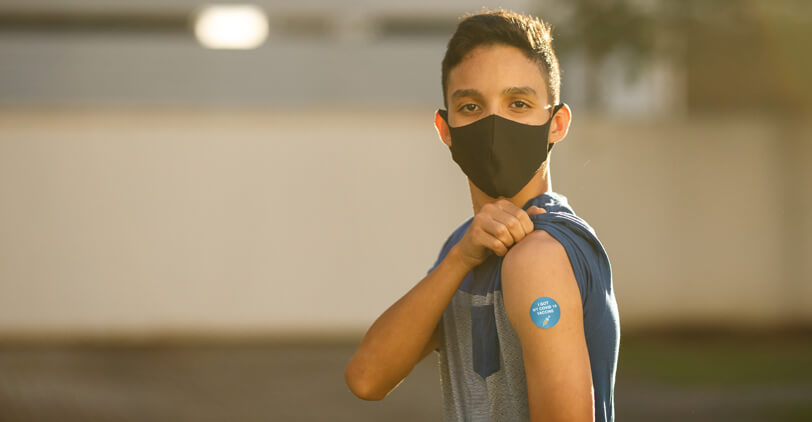 Teenage boy with band aid over vaccine shot location pulling up sleeve wearing mask